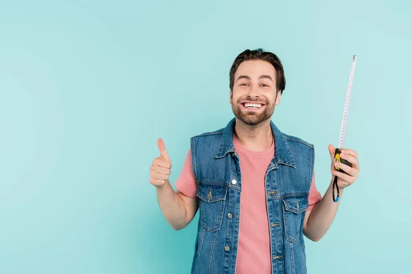 Uomo positivo in gilet di denim che tiene il nastro di misura e mostra come isolato sul blu — Foto stock