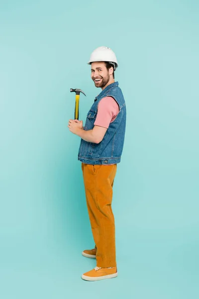 Full length of smiling man in hardhat holding hammer on blue background — Stock Photo