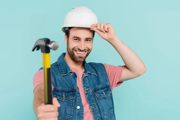 Homem positivo em colete jeans e chapéu duro segurando martelo borrado isolado em azul — Stock Photo