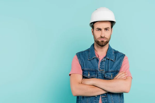 Serious man in hardhat crossing arms and looking at camera isolated on blue — Stock Photo