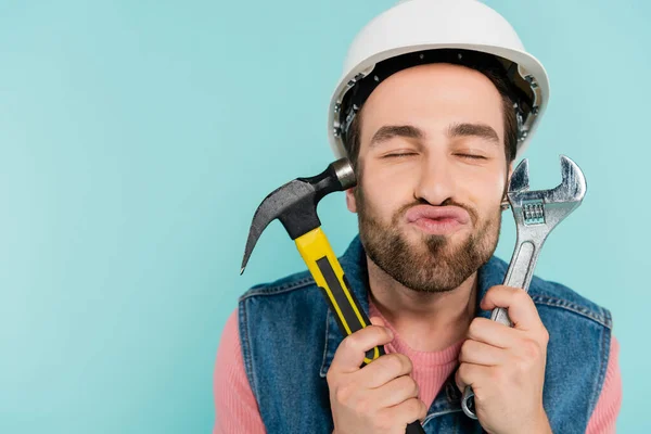 Young man in hardhat pouting lips and holding tools isolated on blue — Stock Photo