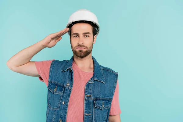 Young man in hardhat saluting and looking at camera isolated on blue — Stock Photo