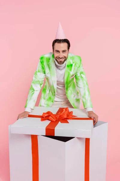 Lächelnder Mann mit Partymütze blickt in die Kamera in der Nähe großer Geschenkschachtel isoliert auf rosa — Stockfoto