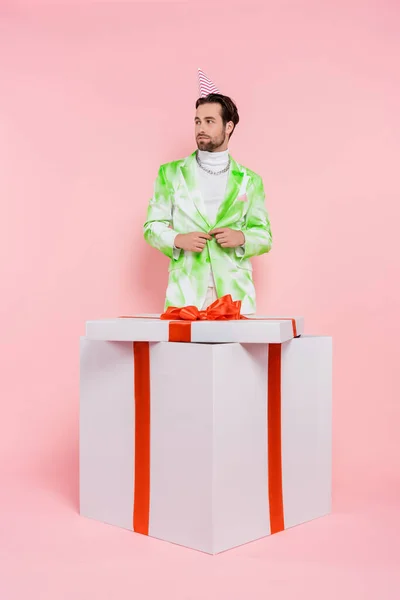 Stylish man in party cap standing near huge present on pink background — Stock Photo