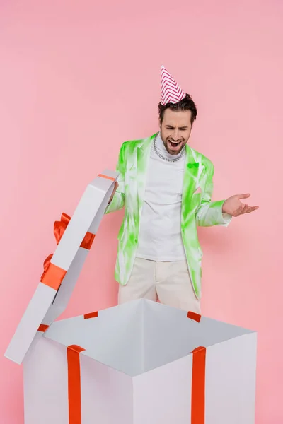 Shocked man in party cap opening big gift box isolated on pink — Stock Photo