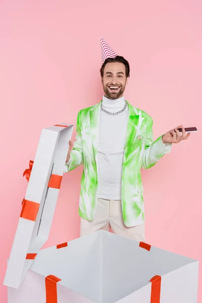 Uomo sorridente nel cappuccio del partito che tiene smartphone vicino alla grande scatola regalo isolata sul rosa — Foto stock