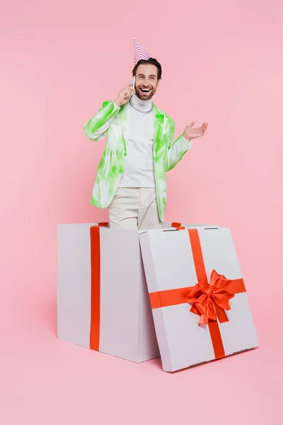 Uomo positivo in berretto partito parlando sul cellulare mentre in piedi in scatola regalo su sfondo rosa — Foto stock
