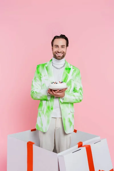 Uomo positivo in cappuccio partito tenendo torta di compleanno mentre in piedi in scatola regalo isolato su rosa — Foto stock