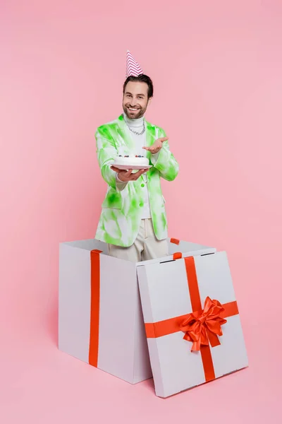 Positiver Mann mit Partymütze zeigt auf Geburtstagstorte in Geschenkbox auf rosa Hintergrund — Stockfoto
