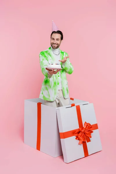 Positive man in party cap pointing at birthday cake in gift box on pink background — Stock Photo