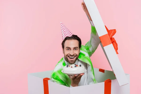 Homem alegre no tampão do partido que prende o bolo de aniversário ao sentar-se na caixa de presente isolada no rosa — Fotografia de Stock