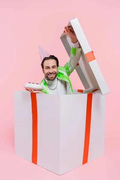 Happy man in party cap holding birthday cake while sitting in gift on pink background — Stock Photo