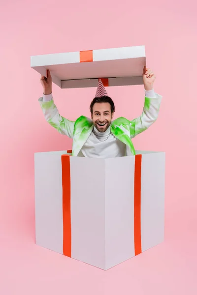 Cheerful man in party cap holding cap of big gift box on pink background — Stock Photo