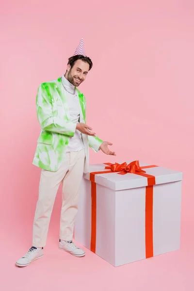 Stylish man in party cap pointing at huge gift on pink background — Stock Photo