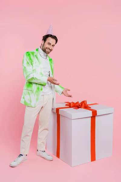 Homme élégant en chapeau de fête pointant vers un énorme cadeau sur fond rose — Photo de stock