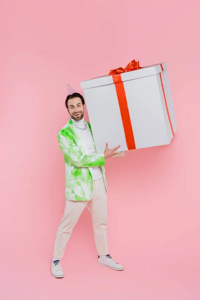 Full length of smiling man in party cap holding big gift box on pink background — Stock Photo