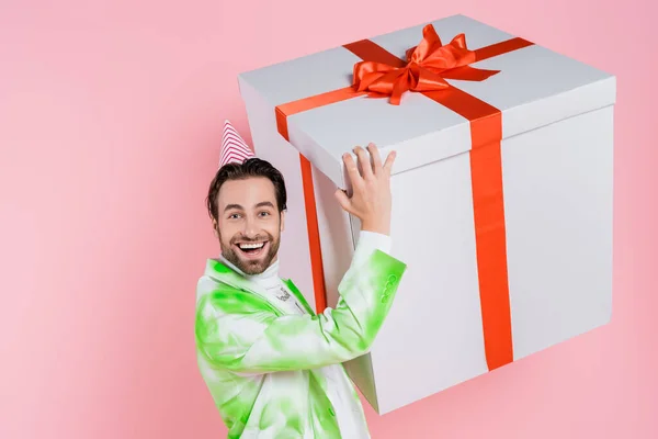 Excited man in party cap and jacket holding big gift box isolated on pink — Stock Photo