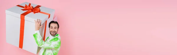 Homem positivo na jaqueta e boné de festa segurando grande presente isolado em rosa, banner — Fotografia de Stock