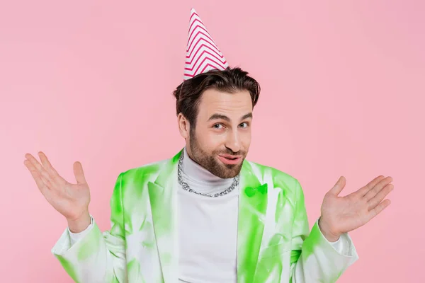 Young man in jacket and party cap gesturing and looking at camera isolated on pink — Stock Photo