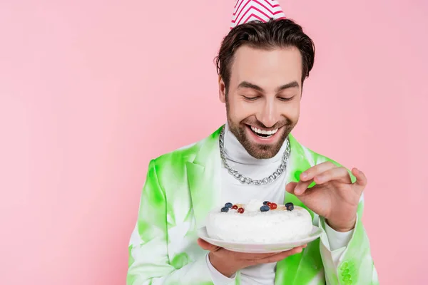 Uomo positivo in tappo partito tenendo torta di compleanno isolato su rosa — Foto stock