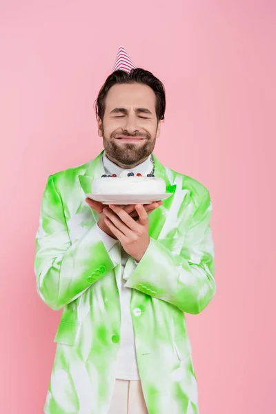 Lächelnder Mann mit Partymütze schließt die Augen und hält Geburtstagstorte isoliert auf rosa — Stockfoto