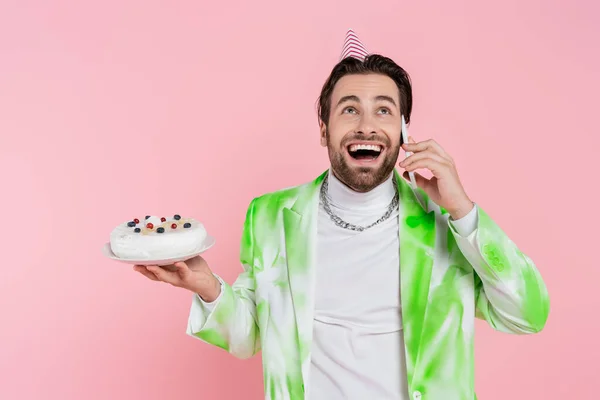 Hombre excitado en la tapa del partido celebración de pastel de cumpleaños y hablando en el teléfono inteligente aislado en rosa - foto de stock
