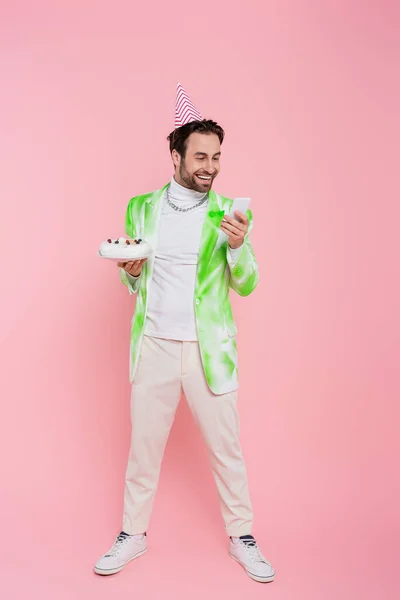 Longitud completa del hombre sonriente en la tapa del partido celebración de pastel de cumpleaños y el uso de teléfono inteligente sobre fondo rosa - foto de stock