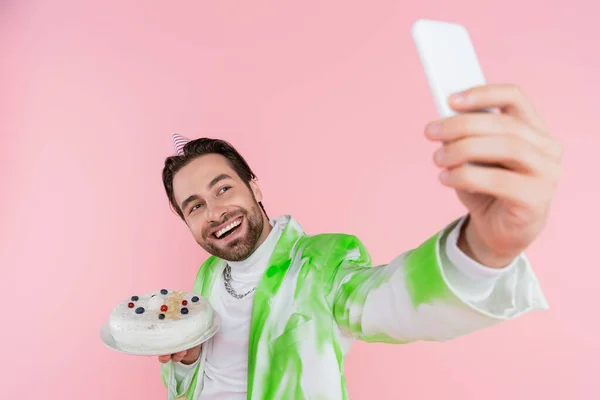 Cheerful man in party cap holding birthday cake and taking selfie on smartphone isolated on pink — Stock Photo