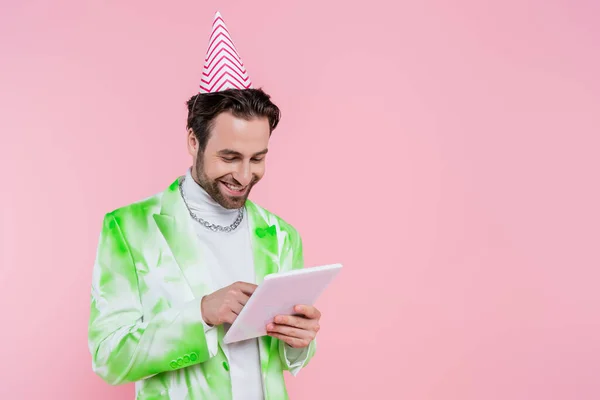 Positiver Mann in Partykappe mit digitalem Tablet isoliert auf rosa — Stockfoto