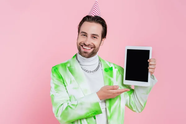 Fröhlicher Mann in Partymütze und Jacke zeigt auf digitales Tablet isoliert auf rosa — Stockfoto