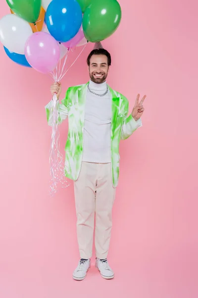 Hombre alegre en gorra de fiesta mostrando señal de paz y sosteniendo globos sobre fondo rosa - foto de stock