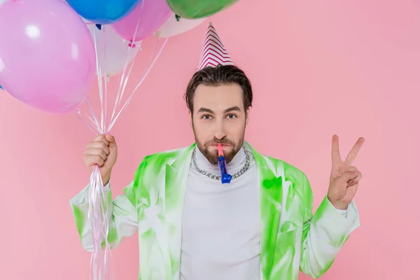 Junger Mann mit Partymütze und Hupe hält Luftballons und zeigt Friedenszeichen isoliert auf rosa — Stockfoto