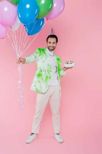 Позитивный человек в кепке для вечеринок держит воздушные шары и торт на розовом фоне — стоковое фото