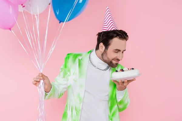 Молодий чоловік у вечірній кепці нюхає торт на день народження і тримає повітряні кулі ізольовані на рожевому — стокове фото
