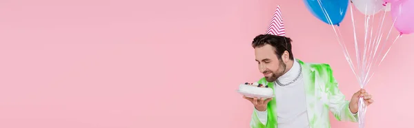 Homme positif dans la casquette de fête sentant gâteau d'anniversaire et ballons isolés sur rose, bannière — Photo de stock
