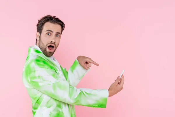 Young man in stylish jacket pointing at smartphone isolated on pink — Stock Photo