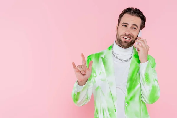 Hombre elegante hablando en el teléfono inteligente y mostrando gesto de roca aislado en rosa - foto de stock