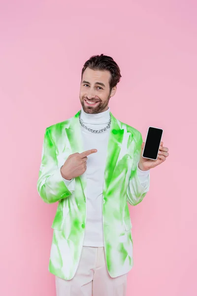 Hombre sonriente con chaqueta apuntando al teléfono inteligente con pantalla en blanco aislado en rosa - foto de stock