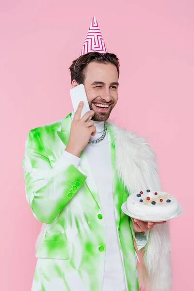 Homme heureux en veste en fourrure et chapeau de fête tenant gâteau d'anniversaire et parler sur smartphone isolé sur rose — Photo de stock