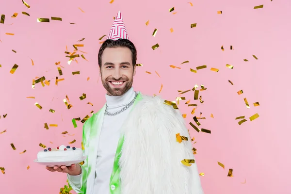 Positive man in furry jacket and party cap holding birthday cake under confetti on pink background — Stock Photo