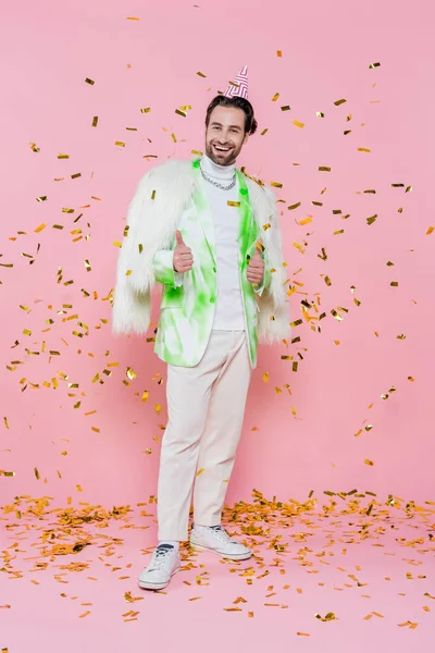 Homem alegre em casaco peludo e tampa da festa mostrando como sob confete no fundo rosa — Fotografia de Stock