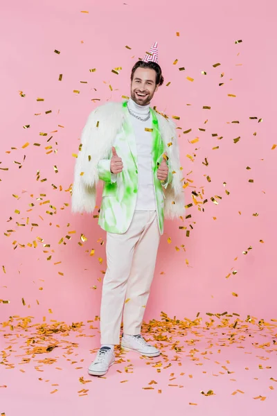 Hombre alegre en chaqueta peluda y gorra de fiesta mostrando como bajo confeti sobre fondo rosa - foto de stock