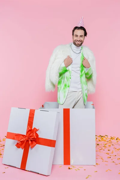 Hombre positivo en gorra de fiesta y chaqueta peluda mirando a la cámara cerca de la caja de regalo enorme y confeti sobre fondo rosa - foto de stock