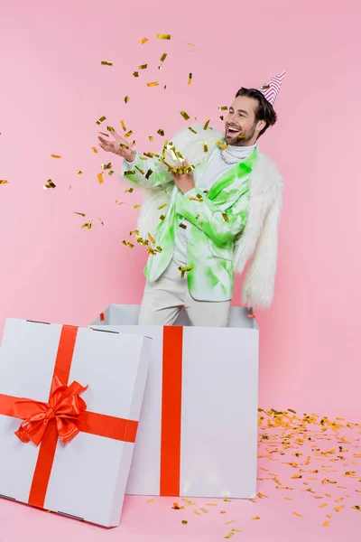 Homme gai en chapeau de fête jetant confettis près de gros présent sur fond rose — Photo de stock