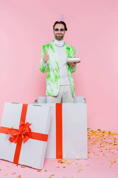 Positiver Mann mit Partymütze und Sonnenbrille zeigt wie und hält Geburtstagstorte in Geschenkbox auf rosa Hintergrund — Stockfoto