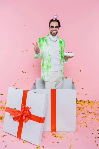 Hombre feliz en gorra de fiesta y gafas de sol mostrando signo de paz y celebración de pastel de cumpleaños en caja de regalo cerca de confeti en rosa - foto de stock