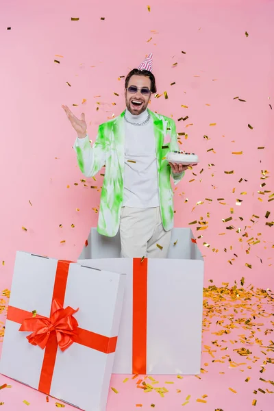 Aufgeregter Mann mit Partymütze hält Geburtstagstorte in Geschenkbox unter Konfetti auf rosa — Stockfoto