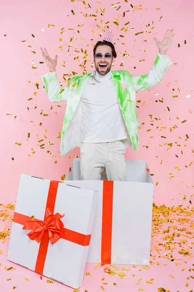 Homme gai dans la casquette de fête debout dans une énorme boîte-cadeau sous confettis sur rose — Photo de stock