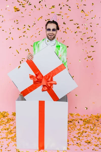 Uomo sorridente in occhiali da sole e copricapo da festa in piedi in confezione regalo sotto i coriandoli su rosa — Foto stock