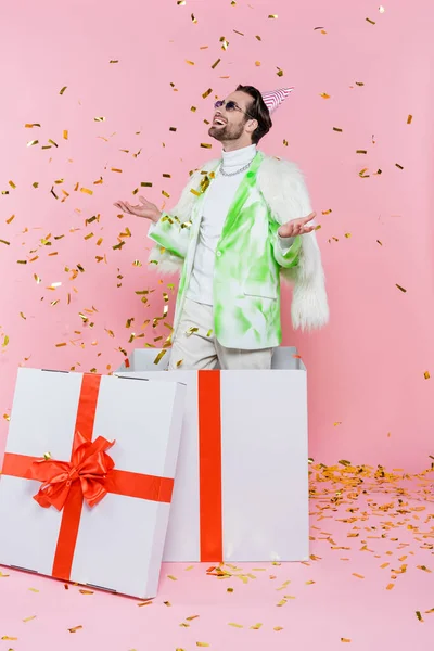 Positive man in party cap standing under confetti near big present on pink background — Stock Photo